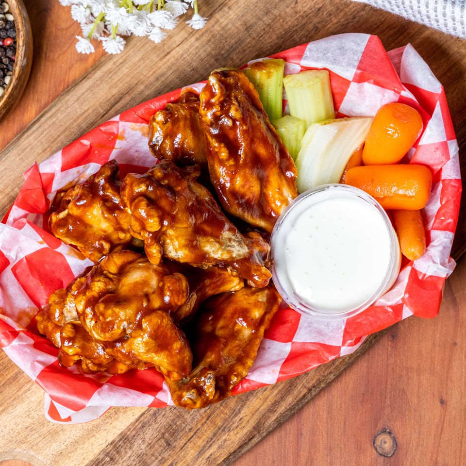 BBQ chicken wings in a red and white checkered paper boat with celery, carrots, and ranch dressing.
