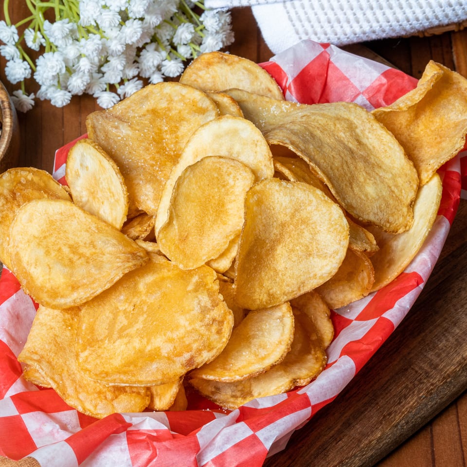Golden potato chips in a red and white checkered basket, with peppercorns and white flowers nearby.