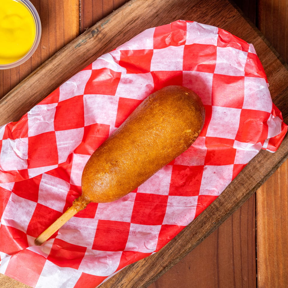 Golden-brown corn dog on red and white checkered paper, with ketchup and mustard cups on a wooden board.