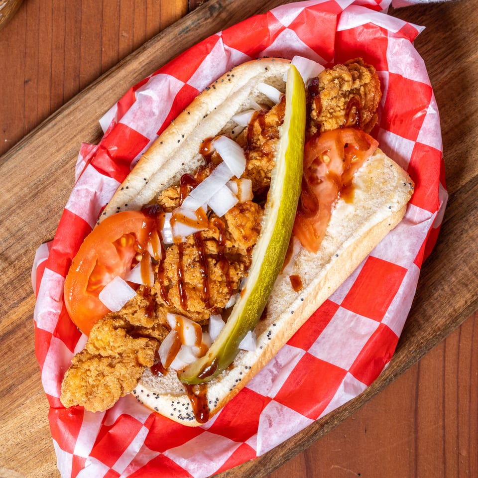 Crispy chicken sandwich with tomatoes, onions, pickle, and BBQ sauce in red checkered paper.