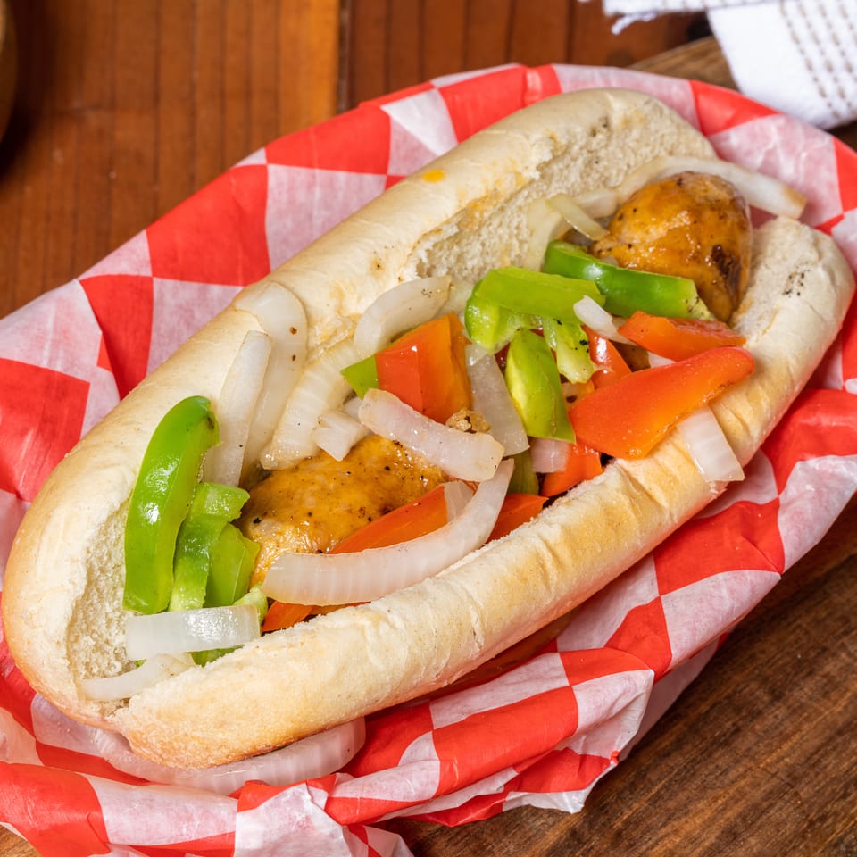Sausage and peppers hero sandwich with onions in a red and white checkered paper boat.