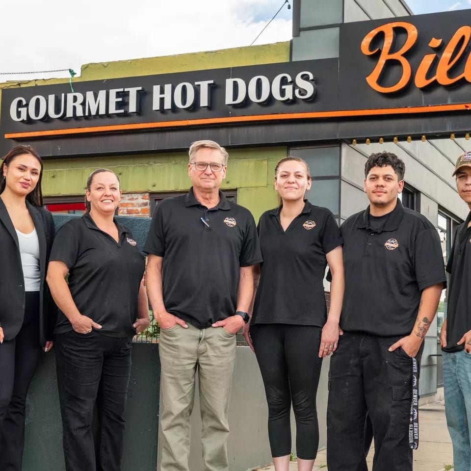 Six Billy's Gourmet Hot Dogs staff members stand smiling outside the restaurant.
