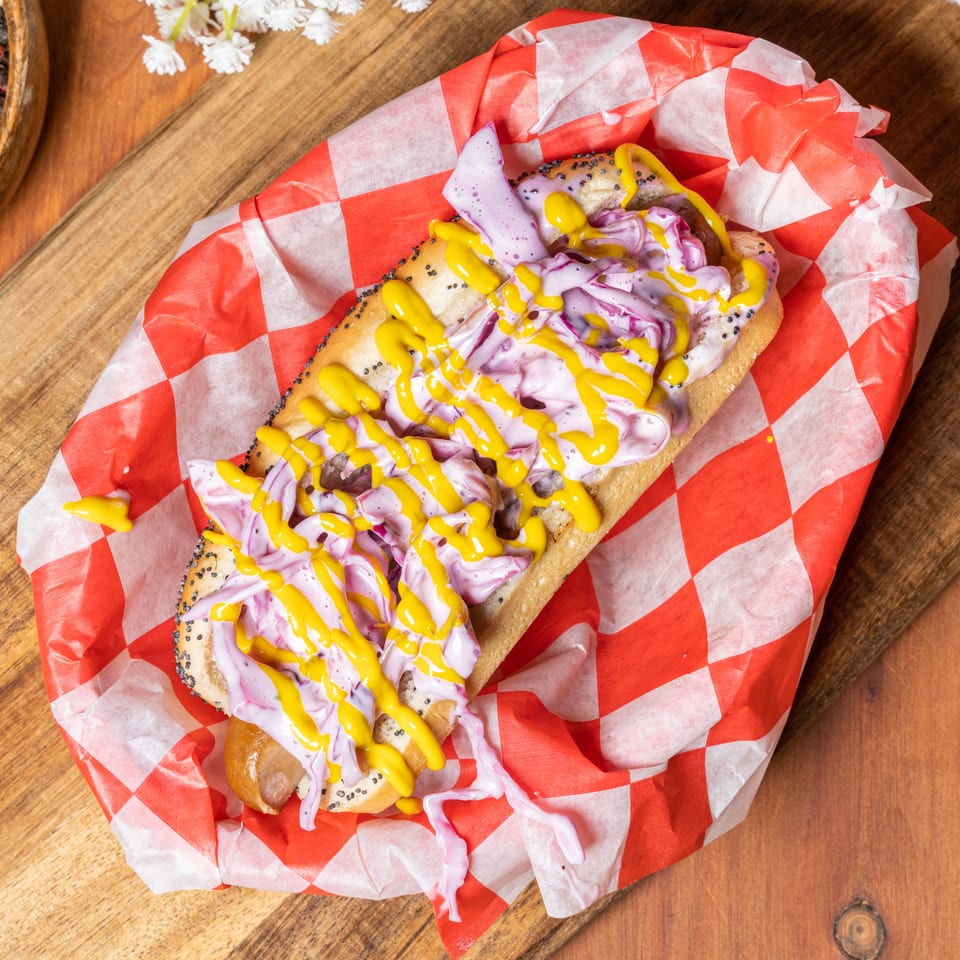 Hot dog with purple coleslaw and yellow mustard in red and white checkered paper on a wooden board.