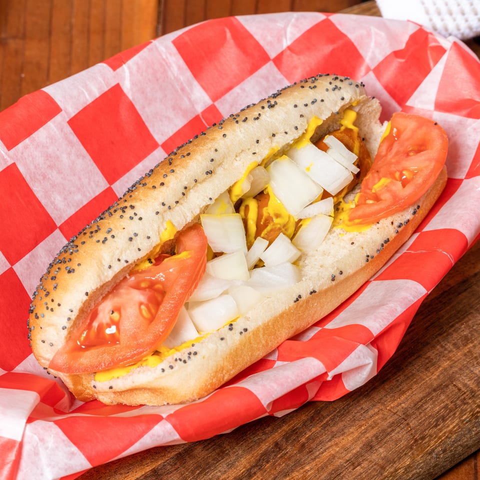 Hot dog with mustard, diced onions, and tomato slices in a poppy seed bun, served in a red checkered paper.