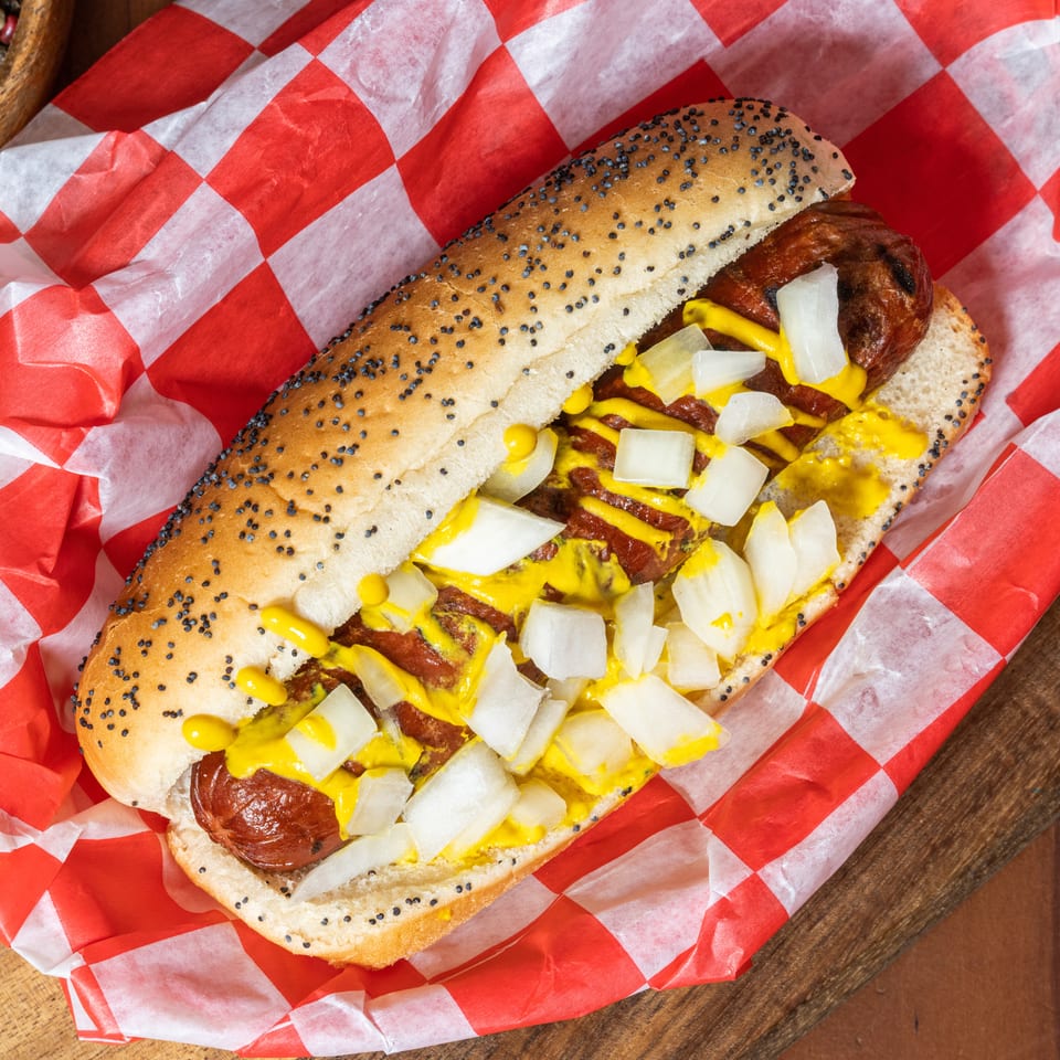 Grilled hot dog with mustard and diced onions on a poppy seed bun, wrapped in checkered paper.