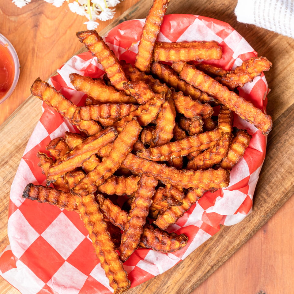 Waffle-cut sweet potato fries in a red and white checkered paper basket with a side of ketchup.