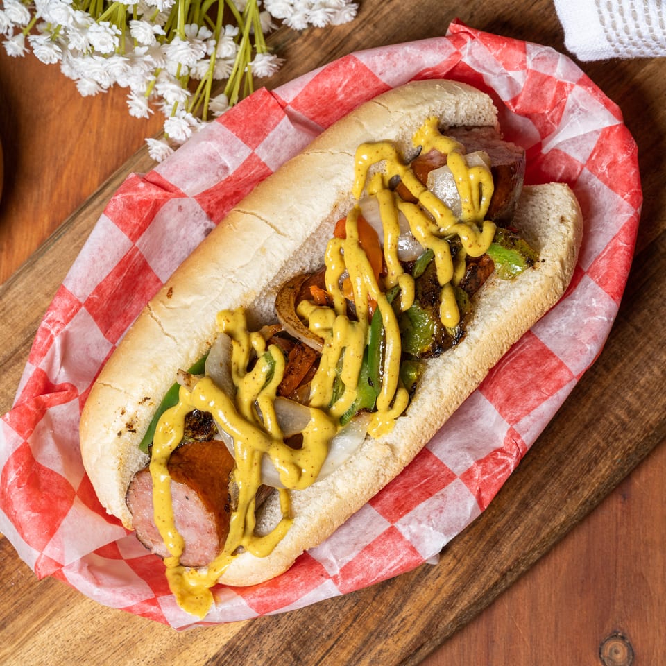 Grilled sausage sandwich with peppers, onions, and mustard in a red checkered wrapper on a wooden board.
